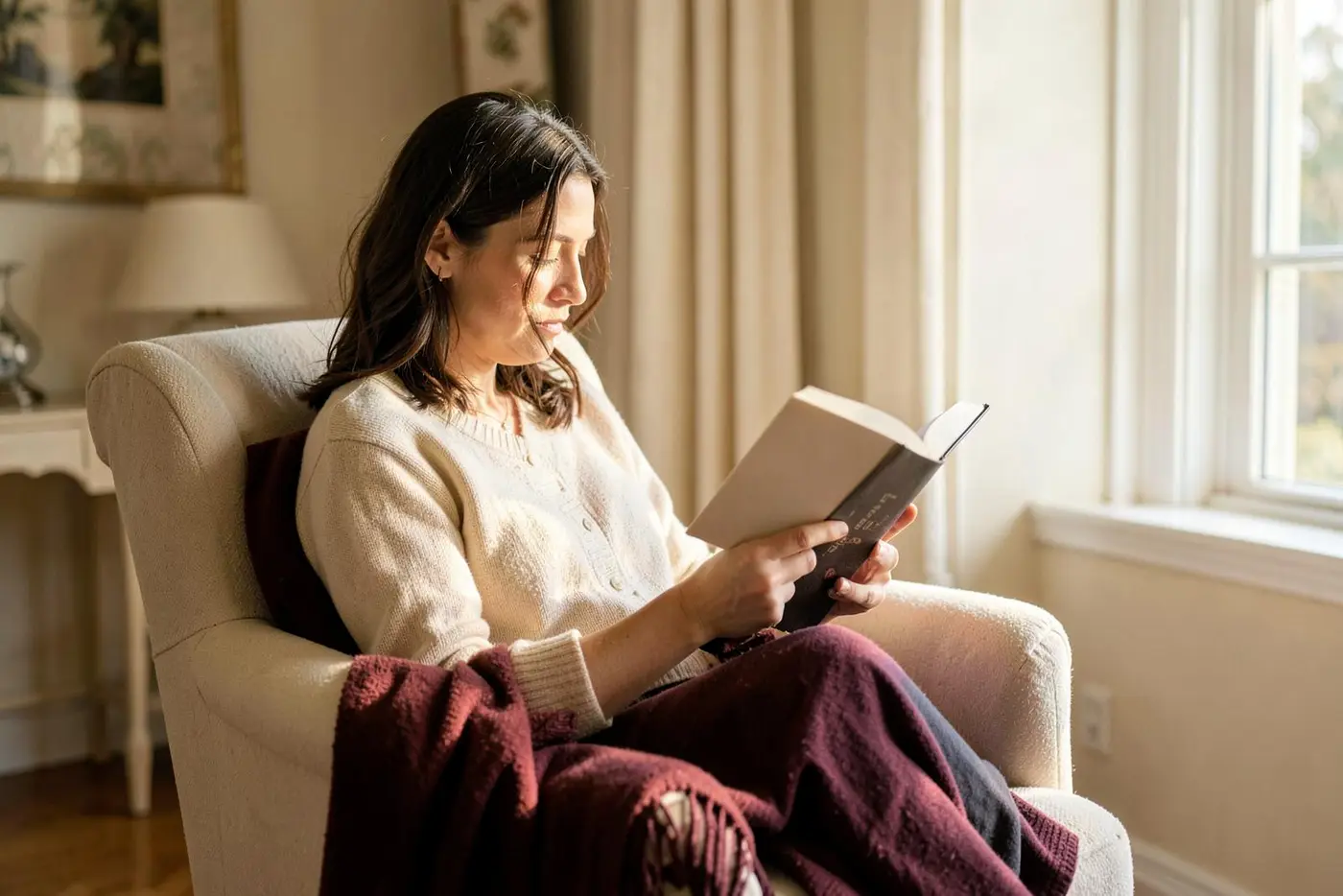 A reader with a hardback book in a comfortable armchair beside a sunlit window